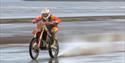 A racing motorcyclist in red racing across the shoreline of a beach