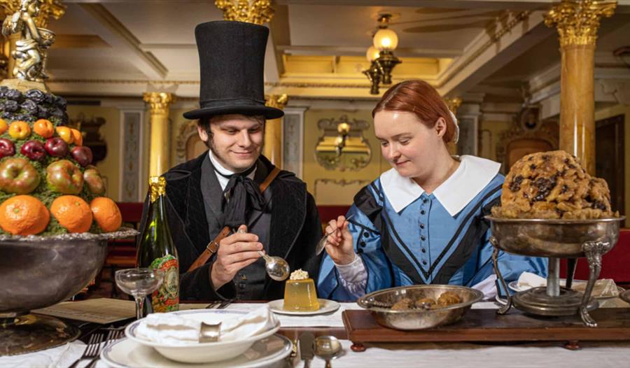 2 people dressed in Victorian costume eating pudding