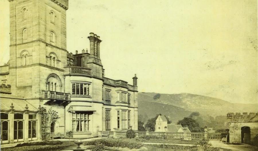 Image is a sepia photograph of Ilam Park, with the historic house and gardens in the foreground, and the Peak District hills behind
