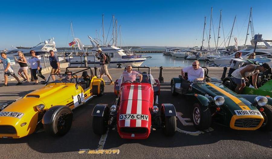 Yellow, red and green roadster style cars lined up at Poole quay with harbour behind them