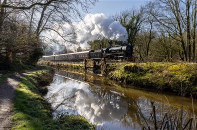 A chugging steam train passes by a river on a sunny day