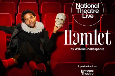 An image of a man sat in a theatre seat wearing black clothing with his left hand which is holding a human skull, raised to be at the same height as h