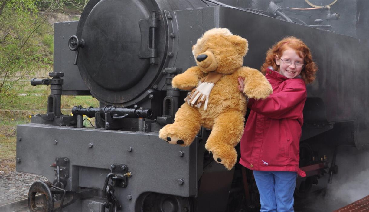 Child holding a teddy bear infront of a steam train
