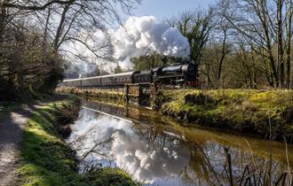 A chugging steam train passes by a river on a sunny day