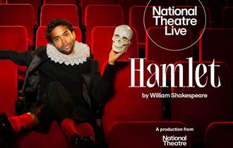 An image of a man sat in a theatre seat wearing black clothing with his left hand which is holding a human skull, raised to be at the same height as h