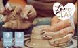 A close-up of two hands covered in clay, as they shape something on the potter's wheel