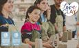 People lined up behind a bench with a lump of clay in hand. Image focuses on girl in pink top and holding clay in her hands, looking at the instructor