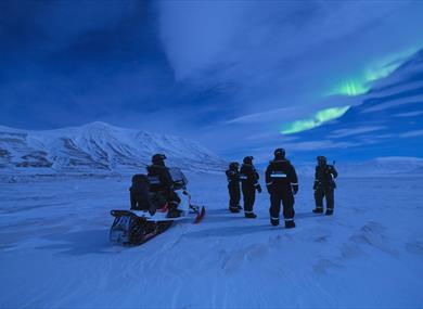 A group of people standing outside with a snowmobile. On the sky its birghr northern lights.