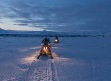 persons driving snowmobiles in a visually blue and snowcovered landscape 