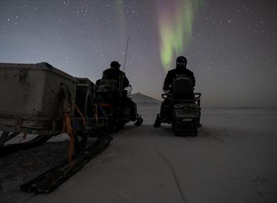 Two people sitting on snowmobiles and looking at the northern lights on the sky
