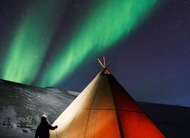 A lighted tent. A person standing outside with headlamp. Green northern ligths on the sky.