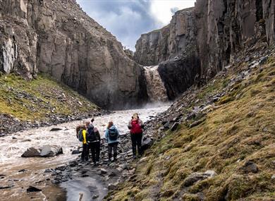 A tour group standing in a valley next to a river. Waterfall in the background.