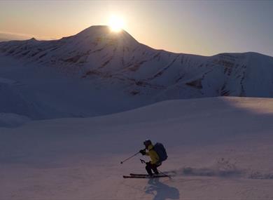 A person skiing down a mountain with powdersnow. Sunset, clear sky and a mountain in the background.