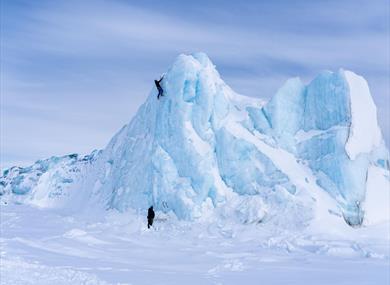 A person climbing a steep ice wall while another stands at the base for safety.