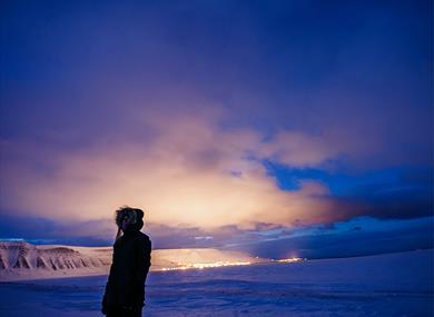 Person standing in front, on a dark winter day with the lights from town in the background 