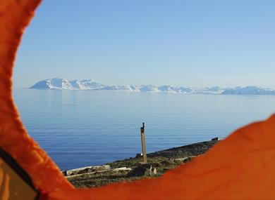 A blue calm fjord with distant snow-covered mountains and a blue sky in the background, seen through the opening of a tent on a hill in the foreground