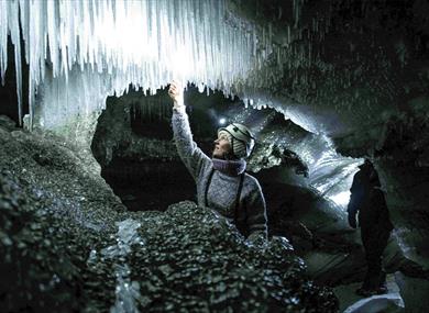 A guide with a bright head lamp and helmet inside an ice cave feeling one of the icicles hanging from the cave roof