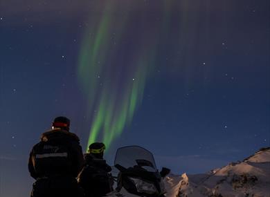 Two persons standing behind a snowmobile and looking up at northern lights in the sky