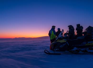 A group of guests and a guide standing behind a snowmobile looking out across a landscape with blue and orange twillight colours in the horizon