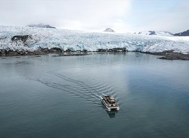 A boat sailing on a fjord in the foreground with a glacier front and mountains surrounding a bay in the background