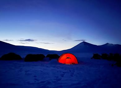 The photo is taken in winter at dusk and shows four tents and some snowmobiles. One of the tents is lit from the inside, glowing red. In the backgroun