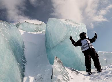 A person stands with his hands in the air in front of a mountain of blue ice.