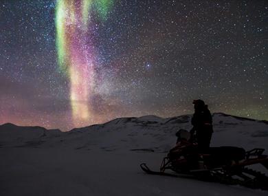A person stands upright on a parked snowmobile, looking at the clear sky adorned with distinct northern lights and stars.