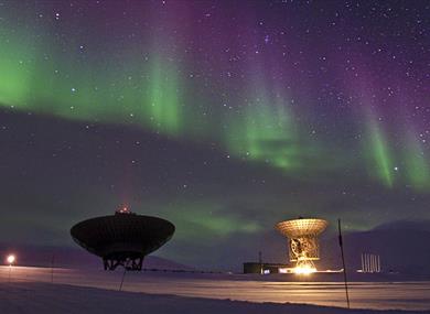 Two radar antennas pointed towards a clear starry sky with northern lights in purple and green colours in the background