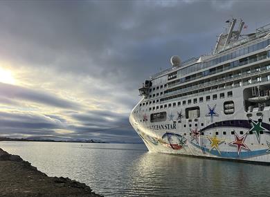 A cruise ship in port inside of a fjord, with an overcast sky and sunshine behind the clouds in the background