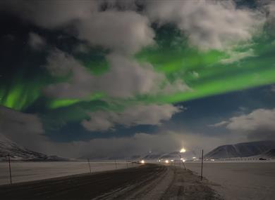 A road winding through a snowy landscape in the foreground, with a dark cloudy sky and northern lights in the background