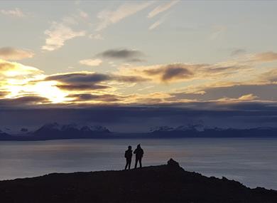 Two persons on top of a mountain in silhouette against a dark fjord and a sunset behind low clouds in the background