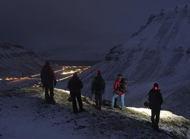 A group of guests with headlamps hiking in dark surroundings, with lights from Longyearbyen in the background