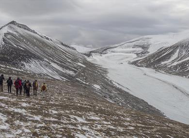 A guide and a group of guests hiking down a mountainside in the foreground with lightly snow-covered mountains and a glacier in the background