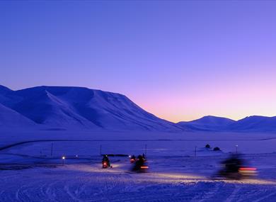 Persons on snowmobiles heading out on a trip with a mountainous landscape covered in blue light in the background