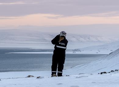 A guest wearing snowmobile equipment standing on a snowy hill scouting out towards a fjord and snowy landscapes in the background
