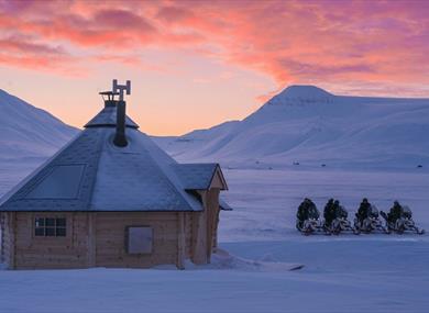 A wilderness cabin and snowmobiles in the snow landscape at sunset.