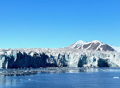 A wide glacier front with a fjord in the foreground and mountains with a clear blue sky in the background