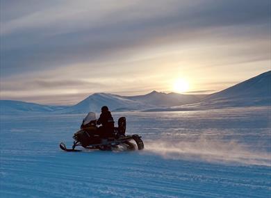Snowmobile driving across a snowy landscape with the sun in the horizon. 