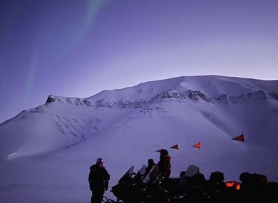 Four snowmobiles parked on a snowy ground. Large mountain in the background. Northern lights in the sky.