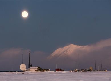 Isfjord Radio in a snowy landscape with mist over the fjord in the background and the moon shining above.