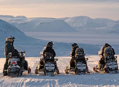 Three guests and a guide on snowmobiles during a stop with snow-covered mountains in the background