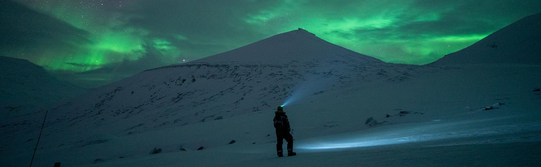 A person with a headlamp in dark snowy surroundings with a mountain and partly cloudy sky with northern lights in the background