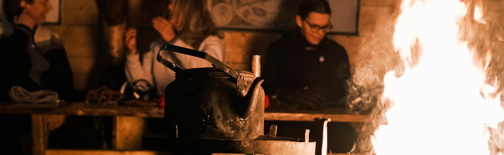 Persons sitting next to a table in the background with a coffee kettle and a fireplace in the foreground