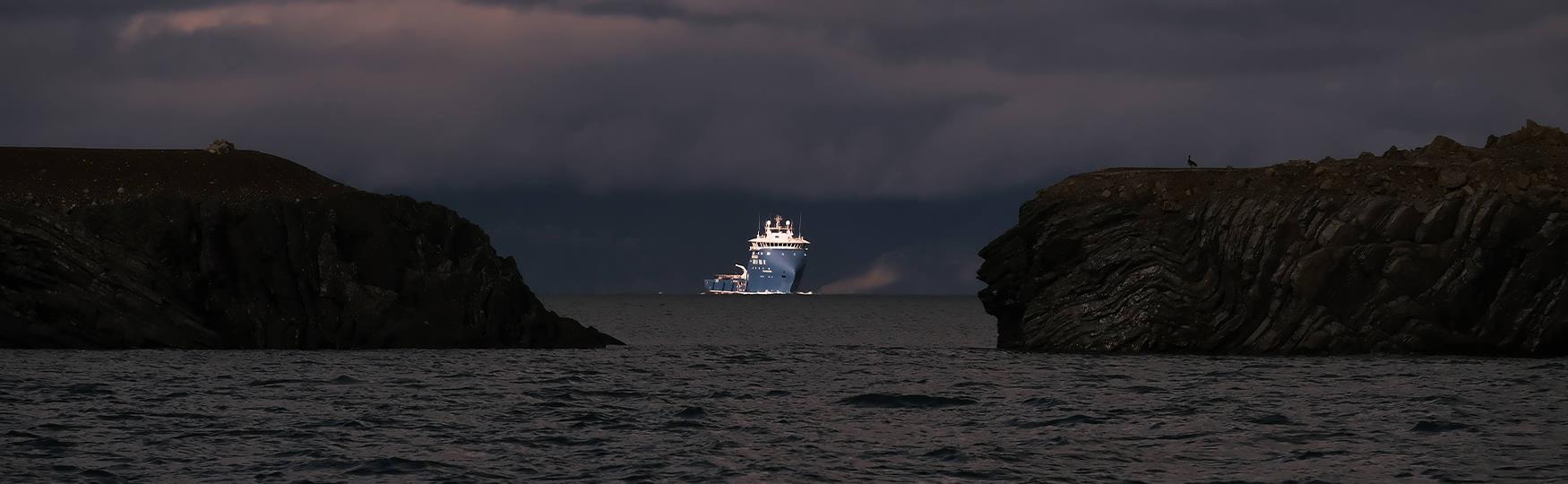 Dark cliffs and a dark fjord in the foreground, with a large ship illuminated by a ray of light amidst dark surroundings on a fjord in the background