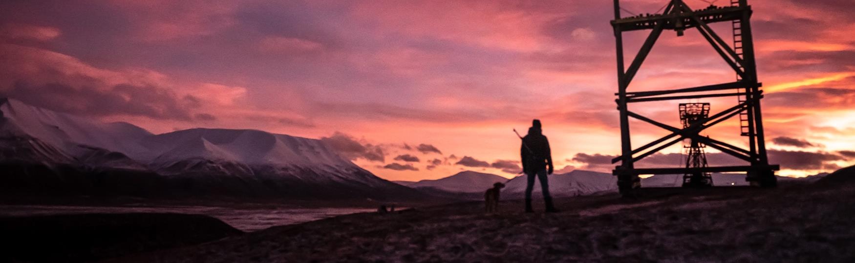 En person og en hund i silhuett ved siden av en taubanebukk med en fargerik himmel og fjell i bakgrunnen