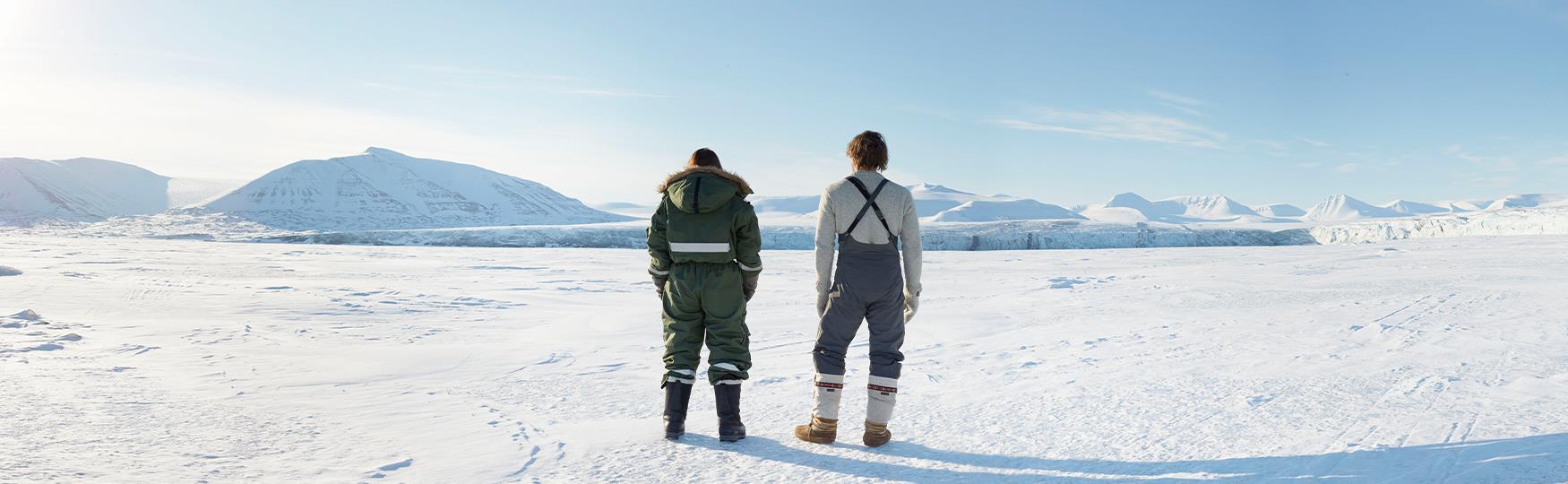 To personer som står ved siden av hverandre i vinterklær i et snødekt landskap. I bakgrunnen ligger en fjellrekke og en isbre.