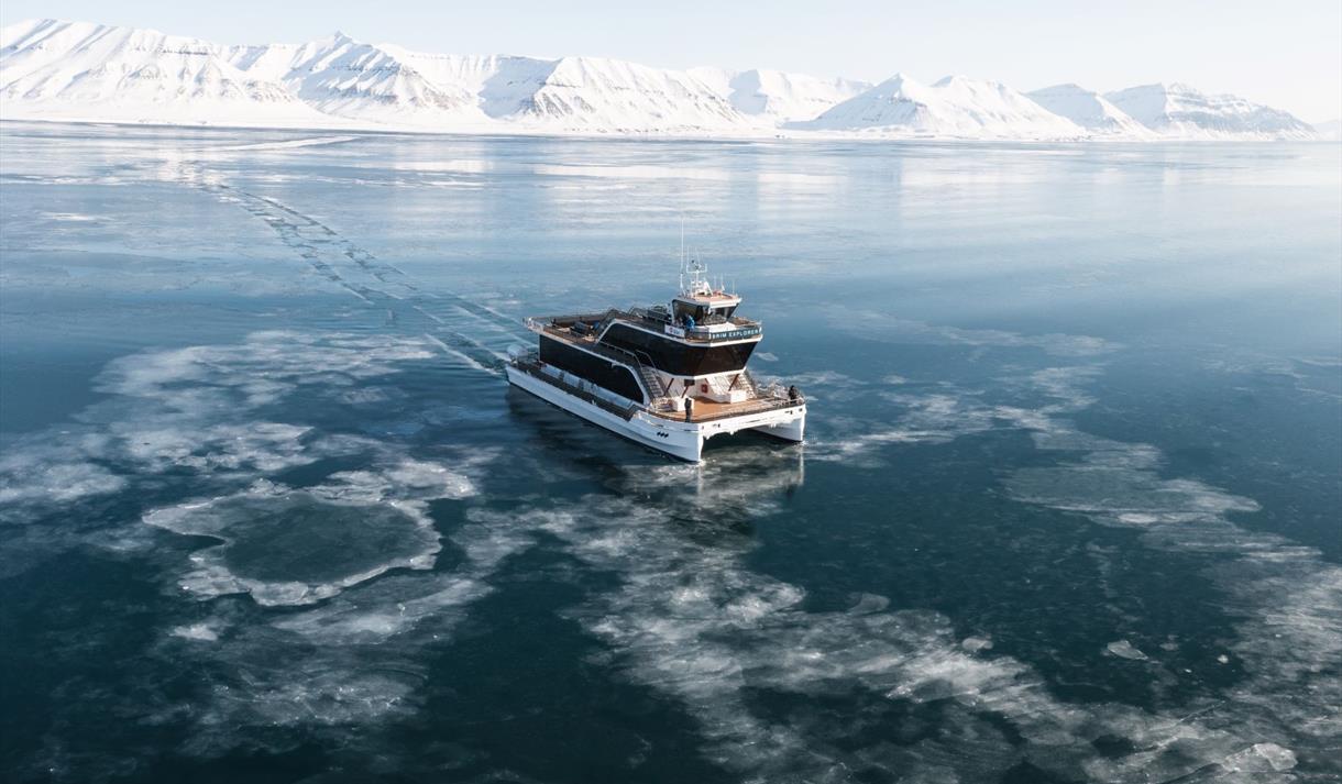 A boat (MS Bard) sailing through thin drift ice on a fjord with mountains in the background