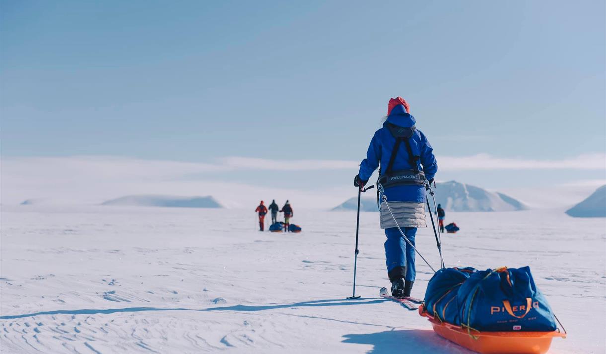 En person på ski som drar en pulk i forgrunnen, med tre personer på ski i bakgrunnen
