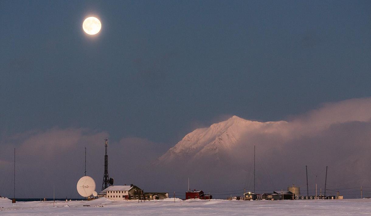 Isfjord Radio i vinterlandskap med tåke over fjorden i bakgrunnen og månen som skinner på himmelen.