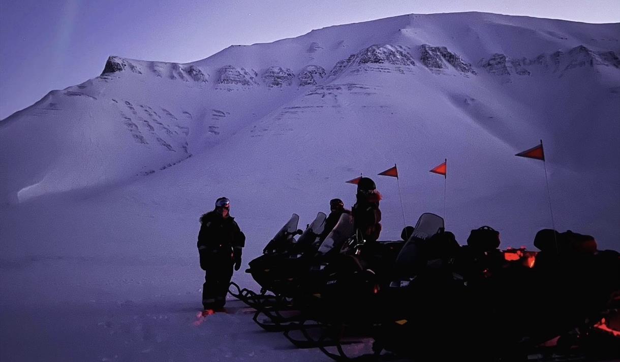 Fire snøscootere står parkert på en slette med et stort fjell i bakgrunnen. Nordlys på himmelen